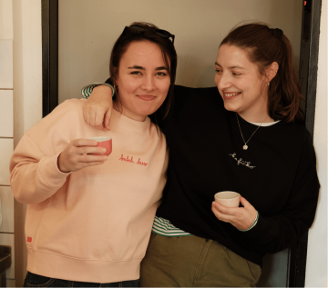 Dvě baristky v legendárních kafíčkových mikinách z Café Jen – broskvové s nápisem „batch brew“ a černé s nápisem „kafíčko“. Pohodlný střih, přirozená atmosféra kavárny.