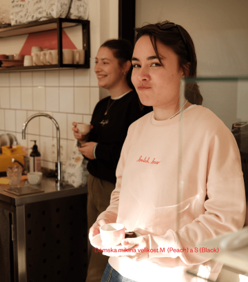 Dvě baristky v legendárních kafíčkových mikinách z Café Jen – broskvové s nápisem „batch brew“ a černé s nápisem „kafíčko“. Pohodlný střih, přirozená atmosféra kavárny.